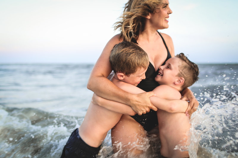 A woman in a swimsuit embraces two laughing boys in the shallow ocean waves, splashing water around. Image by Shayna Hardy Photography Maryland.