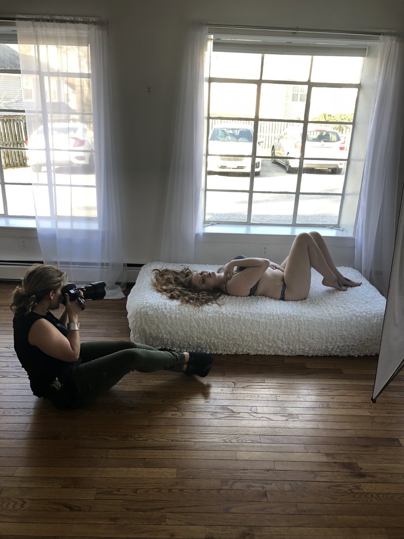 Photographer in green pants taking photos of a woman in a beige bodysuit reclining on a white daybed near an open window with sheer curtains. Image by Shayna Hardy Photography Maryland.