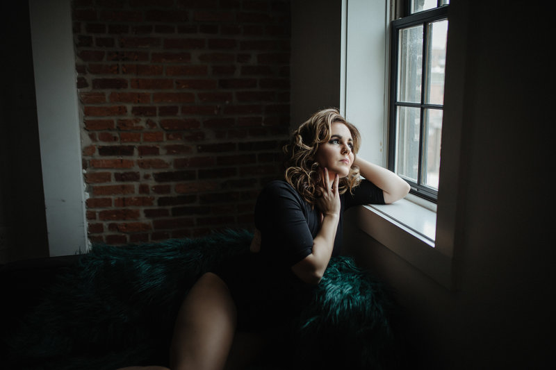 A woman sitting on a fur throw by a window, looking out thoughtfully, in a room with exposed brick walls. Image by Shayna Hardy Photography Maryland.