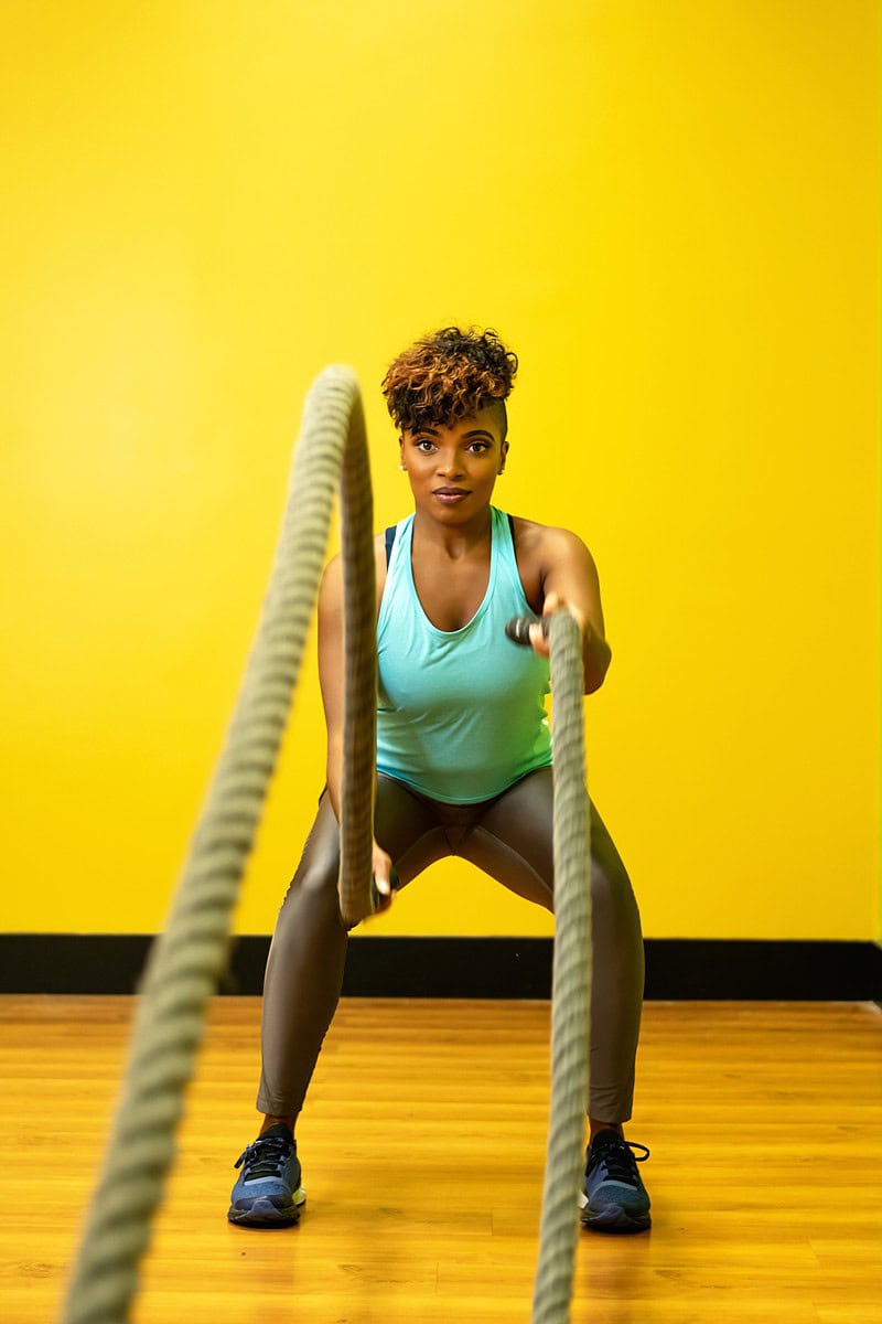 A woman exercises with battle ropes in a gym against a yellow background. She wears a teal tank top and black shorts, focused and determined. Image by Shayna Hardy Photography Maryland.