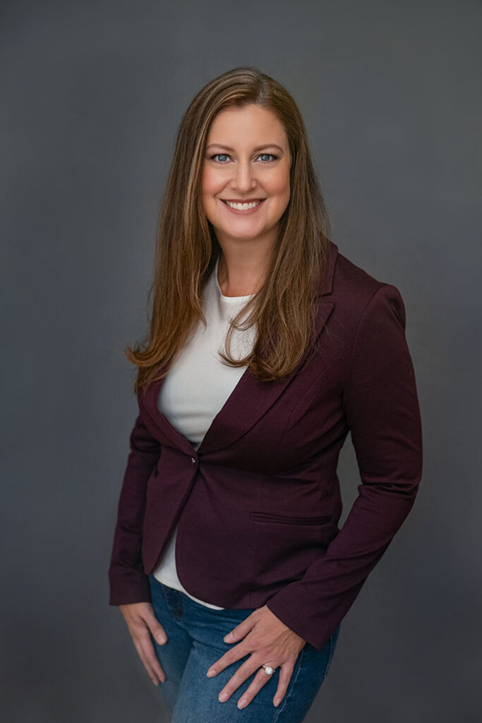 A smiling woman in a burgundy blazer and jeans standing against a gray background. Image by Shayna Hardy Photography Maryland.