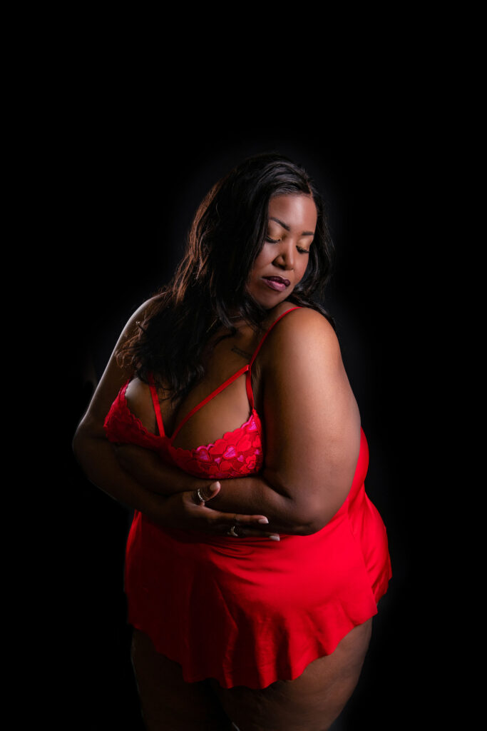 A woman in a red lingerie set poses against a black background, her arms crossed gently over her body and eyes closed. Image by Shayna Hardy Photography Maryland.