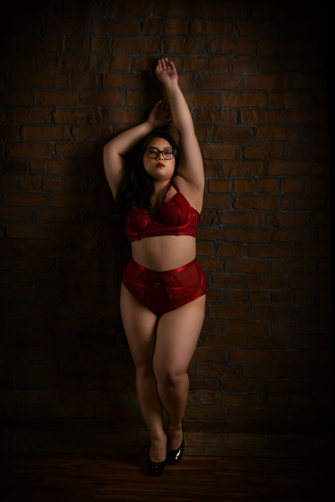 A woman in red lingerie and glasses poses against a brick wall, one arm raised above her head. Image by Shayna Hardy Photography Maryland.