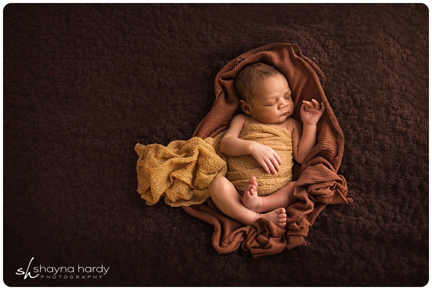 A newborn baby sleeps swaddled in a golden wrap on a brown blanket, with a crumpled golden cloth by their side. Image by Shayna Hardy Photography Maryland.