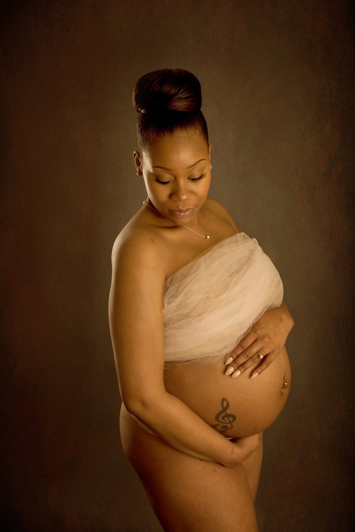 A pregnant woman with a topknot hairstyle, draped in light fabric, holding her belly and looking down, against a textured brown background. Image by Shayna Hardy Photography Maryland.